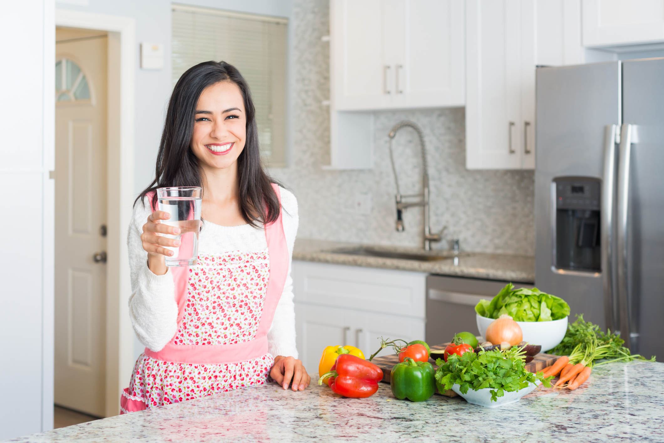 Young Hispanic Women Preparing a Meal Holding A Glass Of Water. Traditional Mexican cuisine.  Salsa, guacamole, limes, cilantro, avocado, carrots, onion,